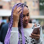 A young woman with vibrant purple braids checks her smartphone in an urban outdoor setting.