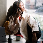 Woman with braids talking on phone in a cafe, sunlight through window.